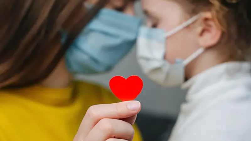 mommy hugs daughter in protective medical masks sitting on bed, holding red heart