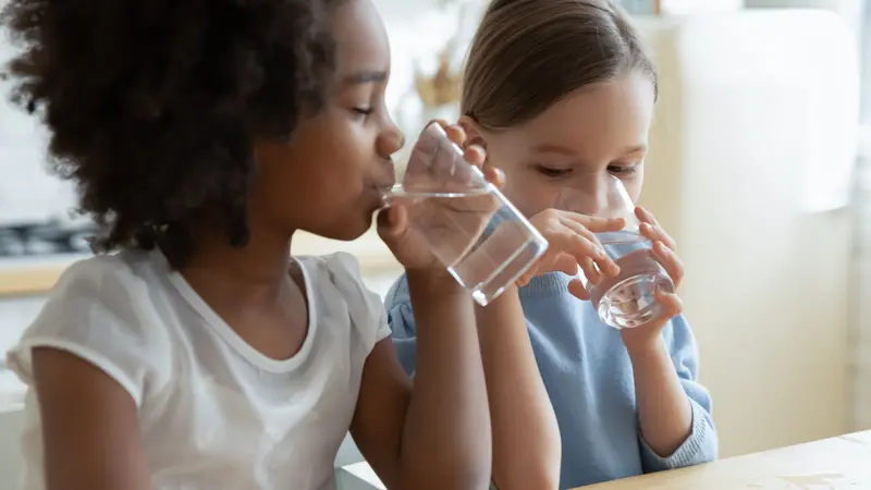 Two multi racial little girls sit at table in kitchen drinking clean water close up image.