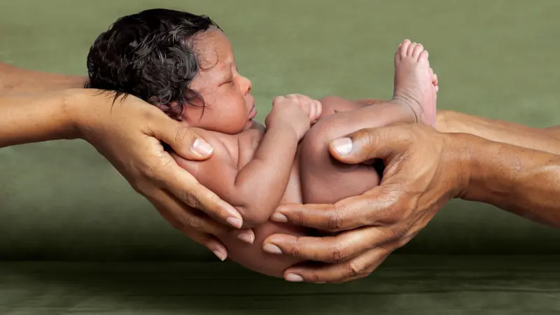 An isolated image of African American parent hands holding their newborn, baby girl in front of a green background.