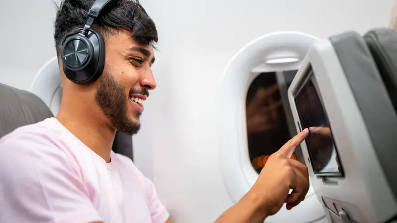 Latin American man in plane cabin using smart device listening to music on headphones.