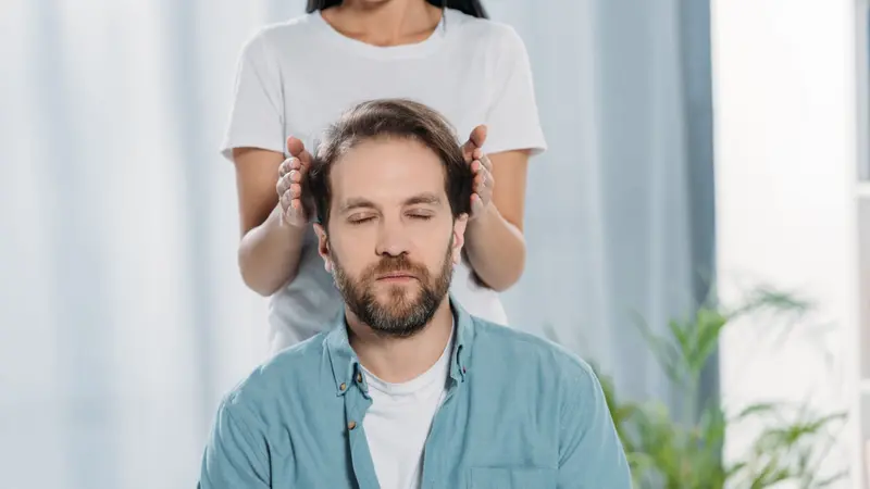 Bearded man with closed eyes sitting and receiving reiki treatment from young female healer