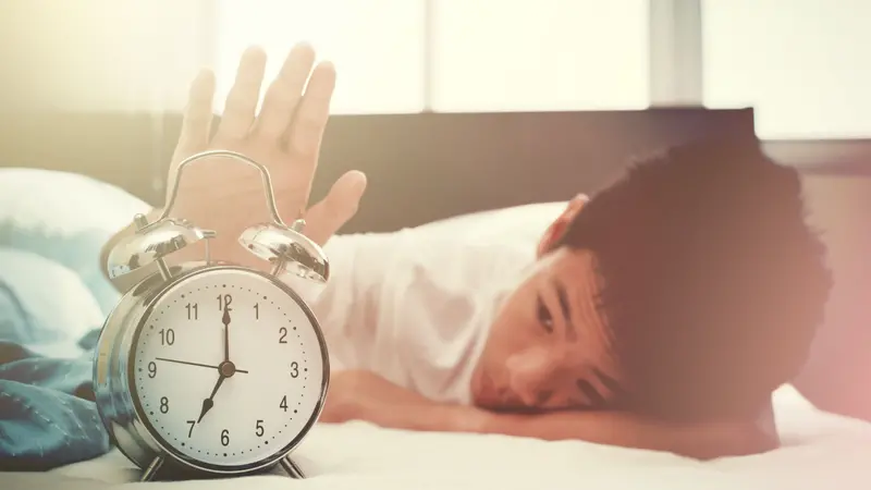 Alarm clock and sleepy teenage boy with hand reaching out for alarm clock